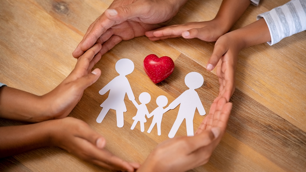Close up hands of father, mother and daughters protecting family paper cutout with red heart. Hands of family with paper cutting and red heart shaped symbol stone. Unity, insurance and love concept.